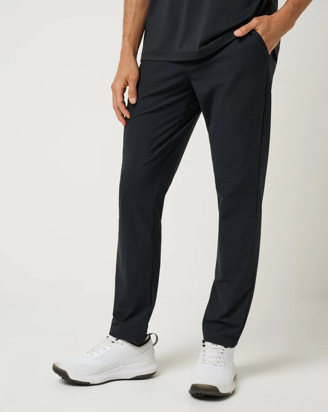A person wearing slim black athletic TravisMathew pants with side pockets, paired with white sneakers, shown from the waist down against a light gray studio background.