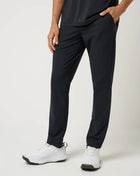 A person wearing slim black athletic TravisMathew pants with side pockets, paired with white sneakers, shown from the waist down against a light gray studio background.