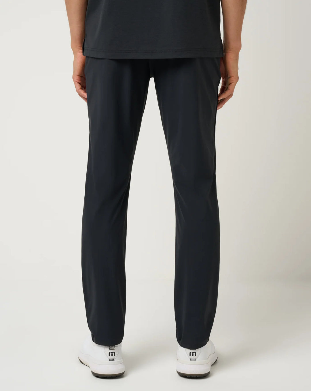A person shown from the back wearing TravisMathew gear including black athletic pants and white athletic shoes against a light gray studio background.