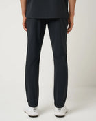 A person shown from the back wearing TravisMathew gear including black athletic pants and white athletic shoes against a light gray studio background.
