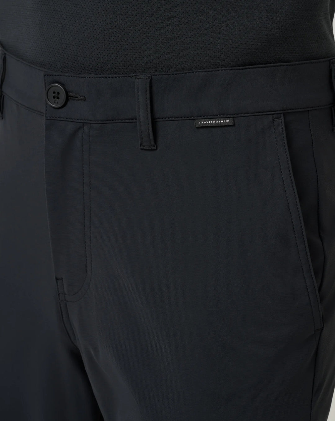 Close-up of dark charcoal gray performance trousers showing the waistband, button closure, belt loops, front pocket, and a small black TravisMathew label.
