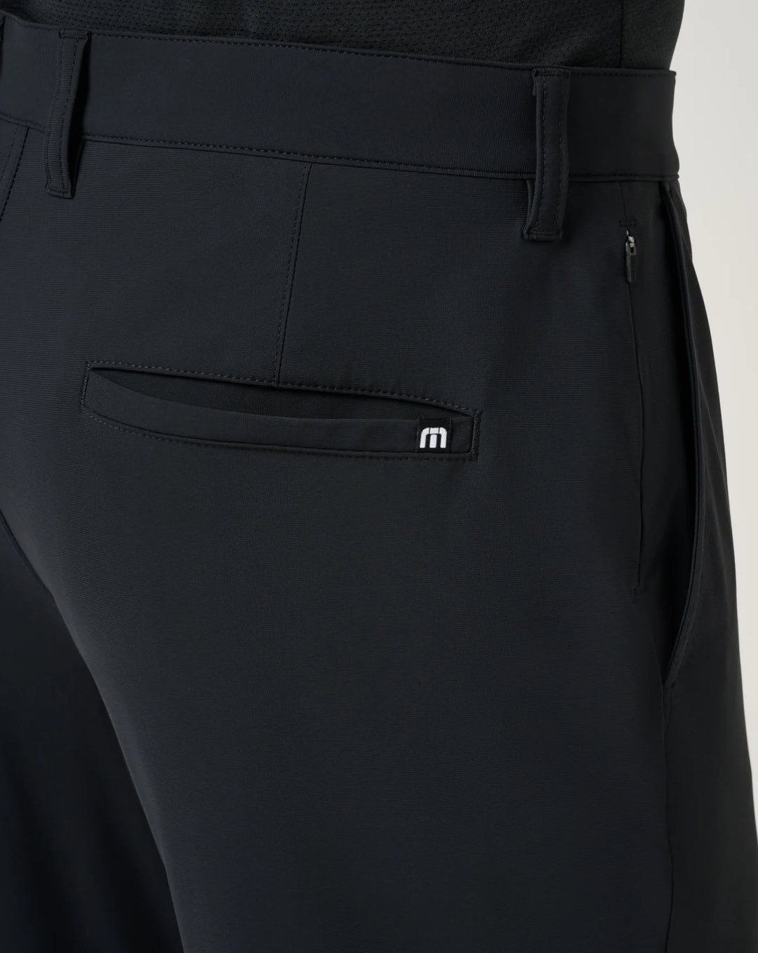 Close-up of black tailored trousers showing the back waistband, belt loops, side zip pocket, and a rear patch pocket with a small white TravisMathew embroidered logo.