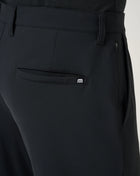 Close-up of black tailored trousers showing the back waistband, belt loops, side zip pocket, and a rear patch pocket with a small white TravisMathew embroidered logo.